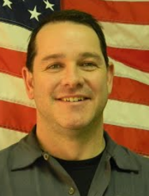 man in front of American flag wearing grey button up shirt